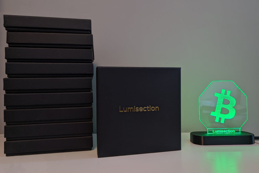 Stack of black boxes with 'Lumisection' branding, next to a glowing green LED sign with Bitcoin symbol.